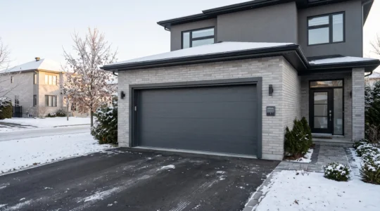 Porte de garage double moderne en acier gris anthracite installée sur une maison unifamiliale de banlieue montréalaise, avec panneau de contrôle automatique visible et contexte hivernal canadien